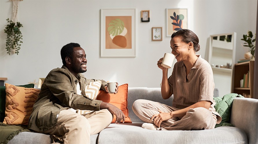 En kvinde og en mand sidder i en sofa, smilende, men med hver sin kop i h�nden.