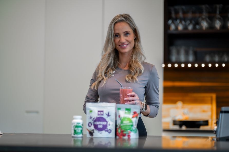 Lea Kessler str i et kkken, hvor hun holder en smoothie, foran produkter fra Healthwell.