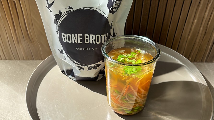 Et glas med nudelsuppe st�r p� et bord sammen med en pose Healthwell Benbouillon Pulver Gr�sfodret.