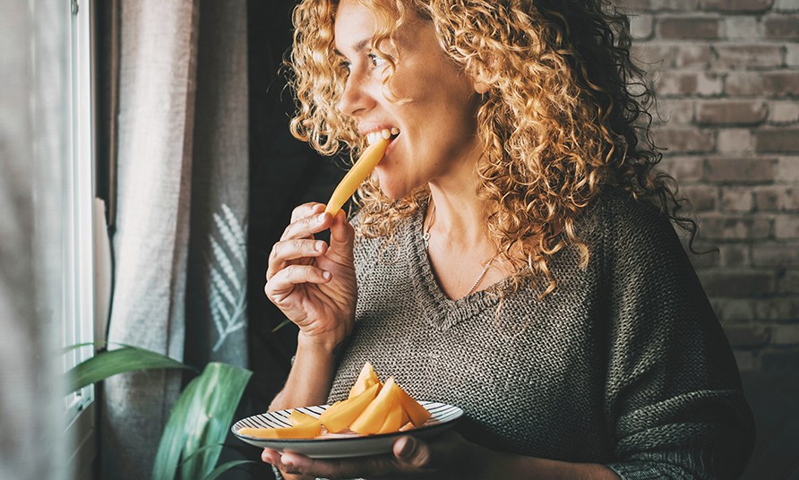 En kvinde str ved et vindue og spiser et stykke frugt.