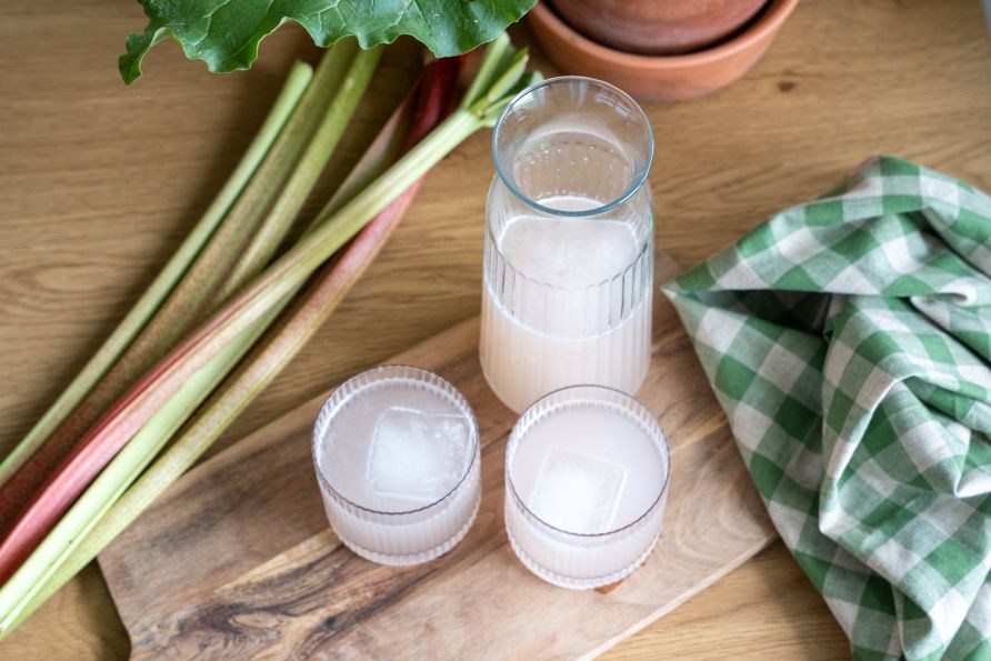 Hjemmelavet rabarbersaft med isterninger serveret i glas og karaffel, med friske rabarberstilke ved siden af p� et tr�br�t.