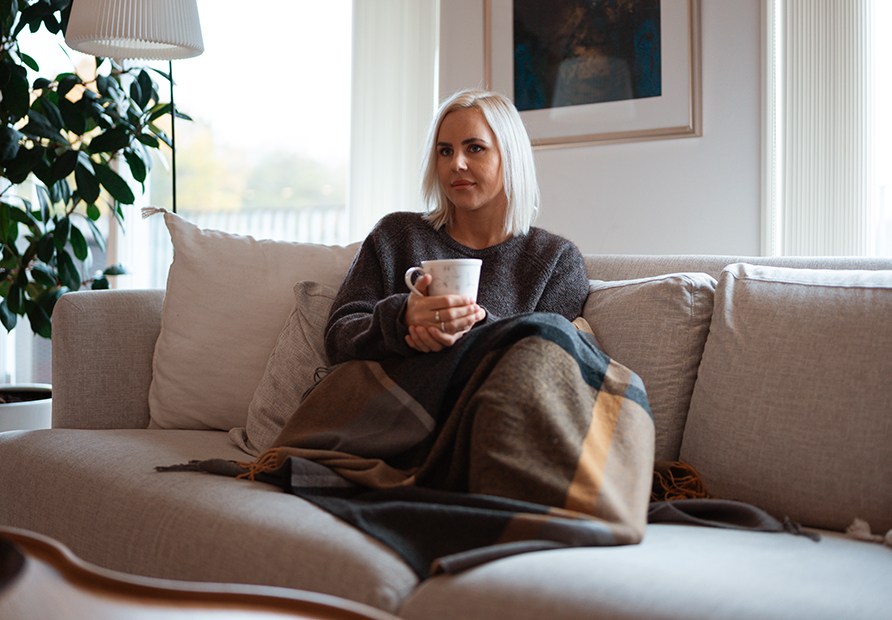 Sundhedscoach Susanna Jungblom sidder sammenkrøbet i en sofa med et tæppe og en kop te. Sundhedscoach Susanna Jungblom sidder sammenkrøbet i en sofa med et tæppe og en kop te.
